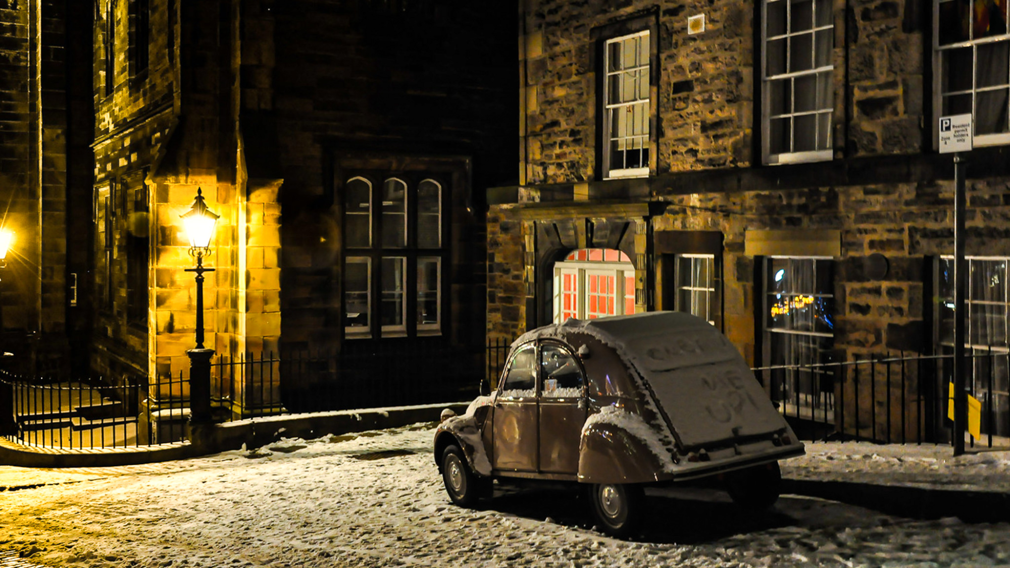 Snow-covered vintage Citroën 2CV on a cobblestone street at night in Edinburgh, Scotland, with “Clean Me Up” written on the rear window under warm streetlight